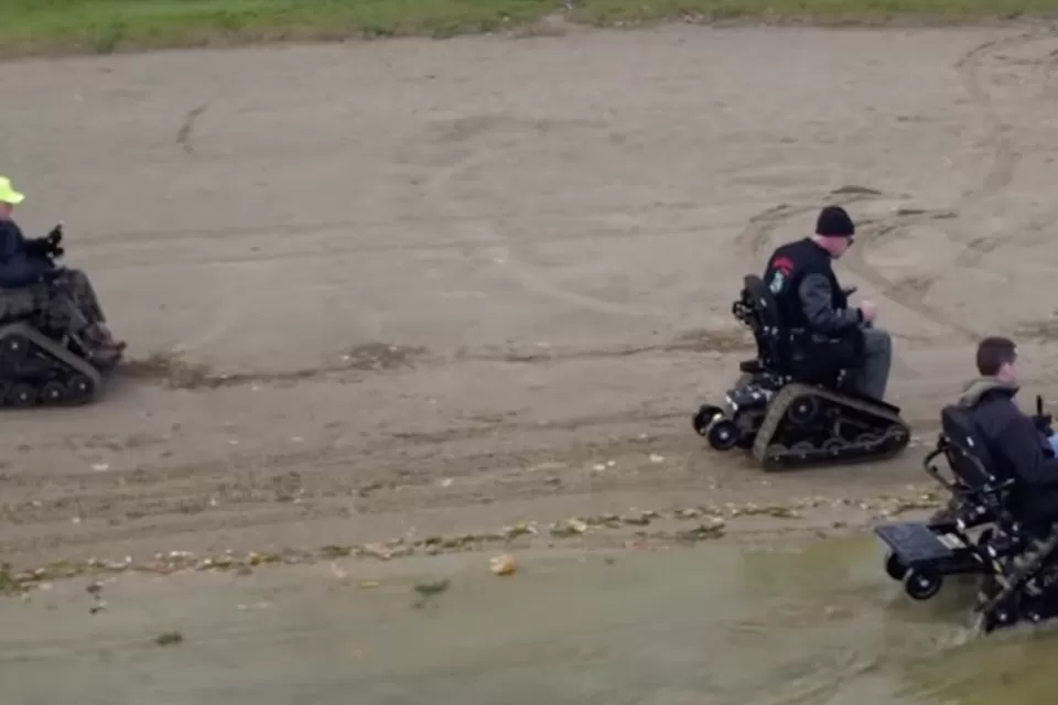 Aerial view of three men using track chair mobility devices on lake beach in Michigan state park.